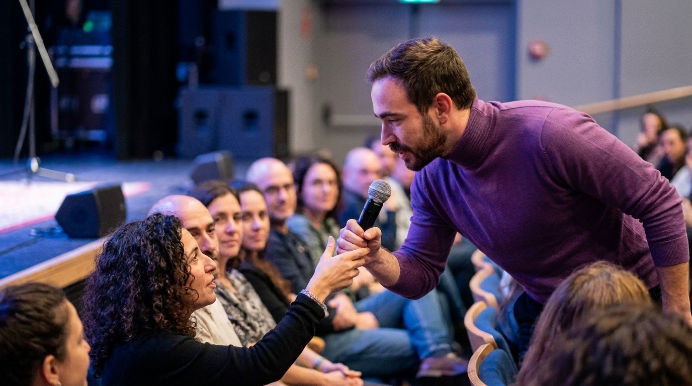 נטע ברסלר מושיט מיקרופון לאורחת באולם בחיפה בעוד הקהל היושב סביבה פורץ בצחוק