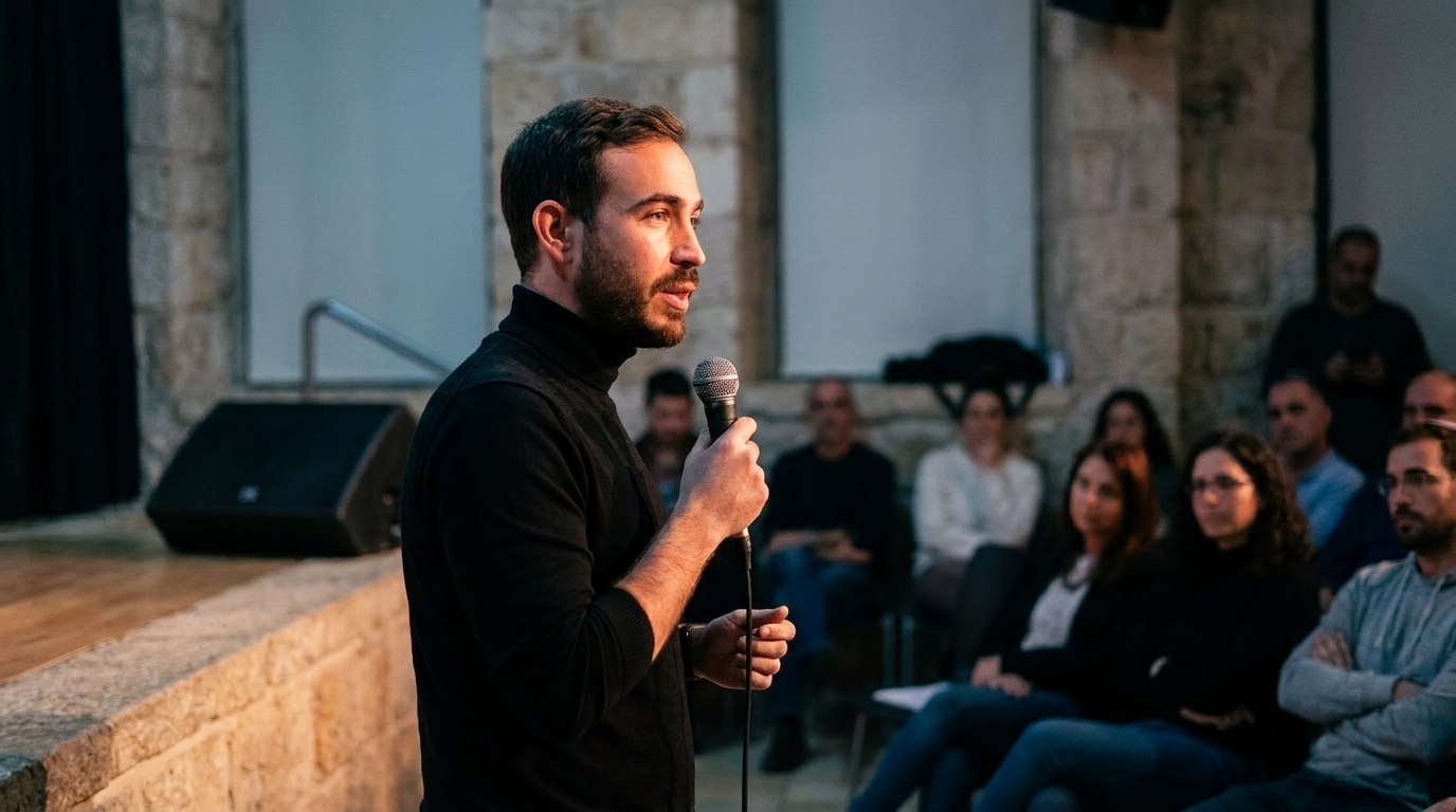 Neta Bresler stands on a low stage in a Jerusalem stone hall, holding a microphone out toward a seated audience.